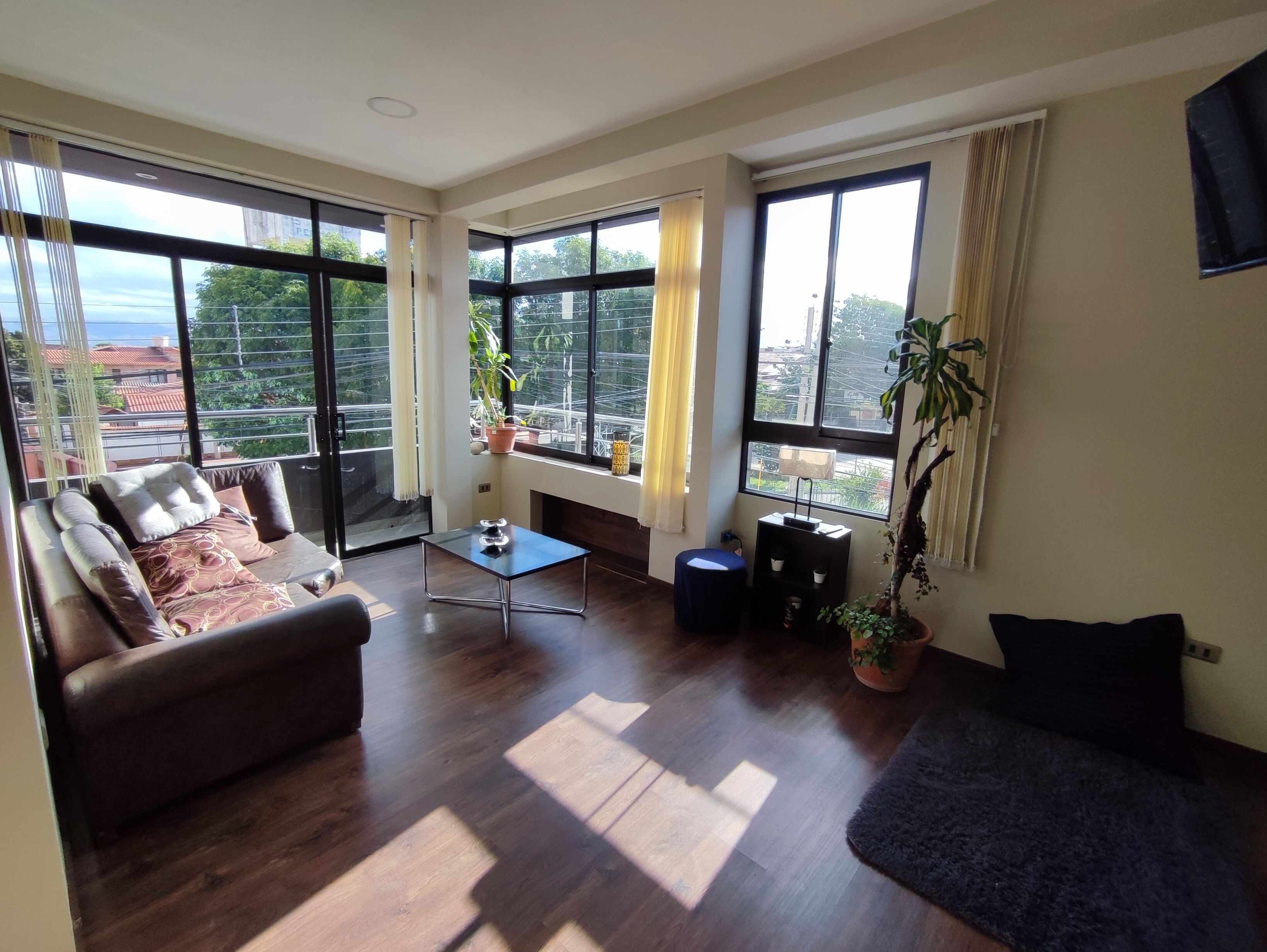 A spacious living room with a sofa, a coffee table, and a blue ottoman. Large windows provide ample natural light and views of the city.