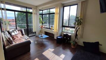 A spacious living room with a sofa, a coffee table, and a blue ottoman. Large windows provide ample natural light and views of the city.