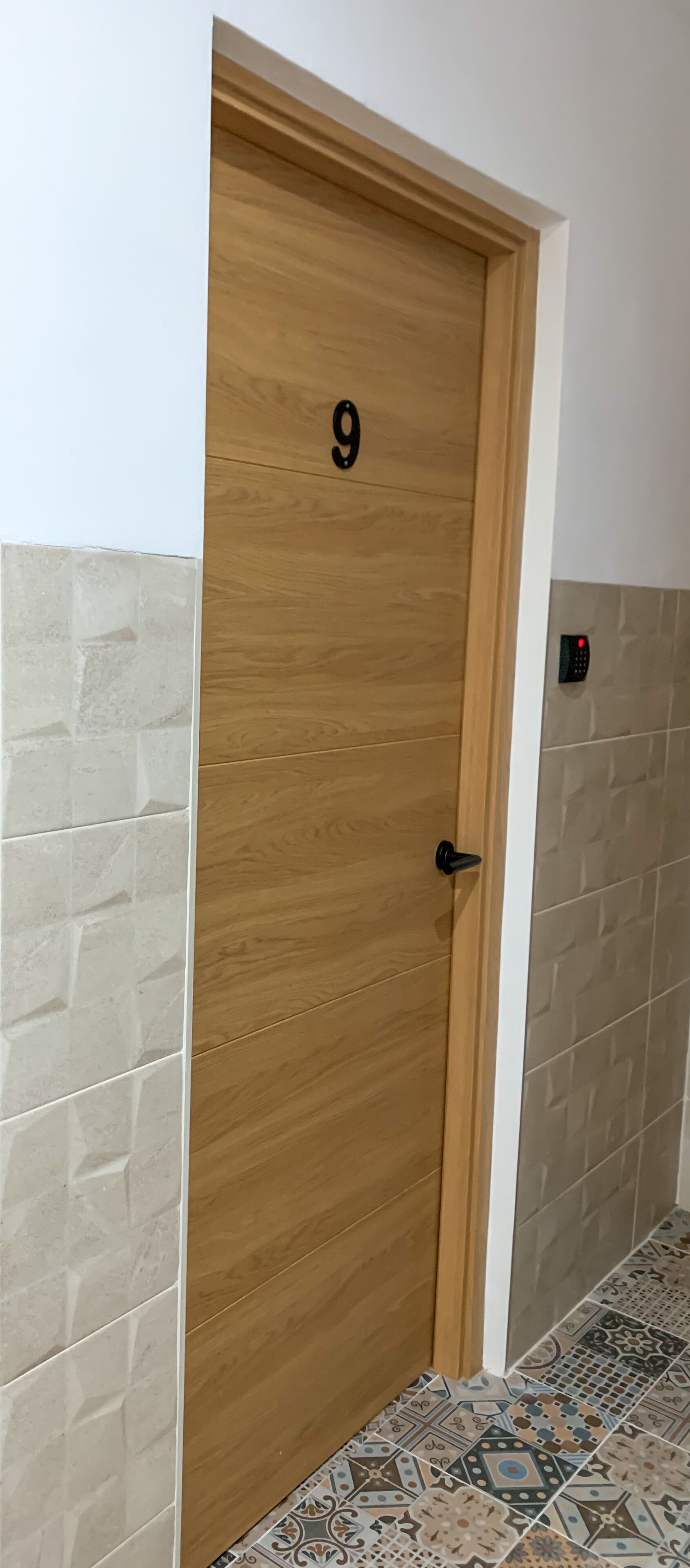 A wooden door with the number 9, a black door handle, and a wall with patterned tiles.