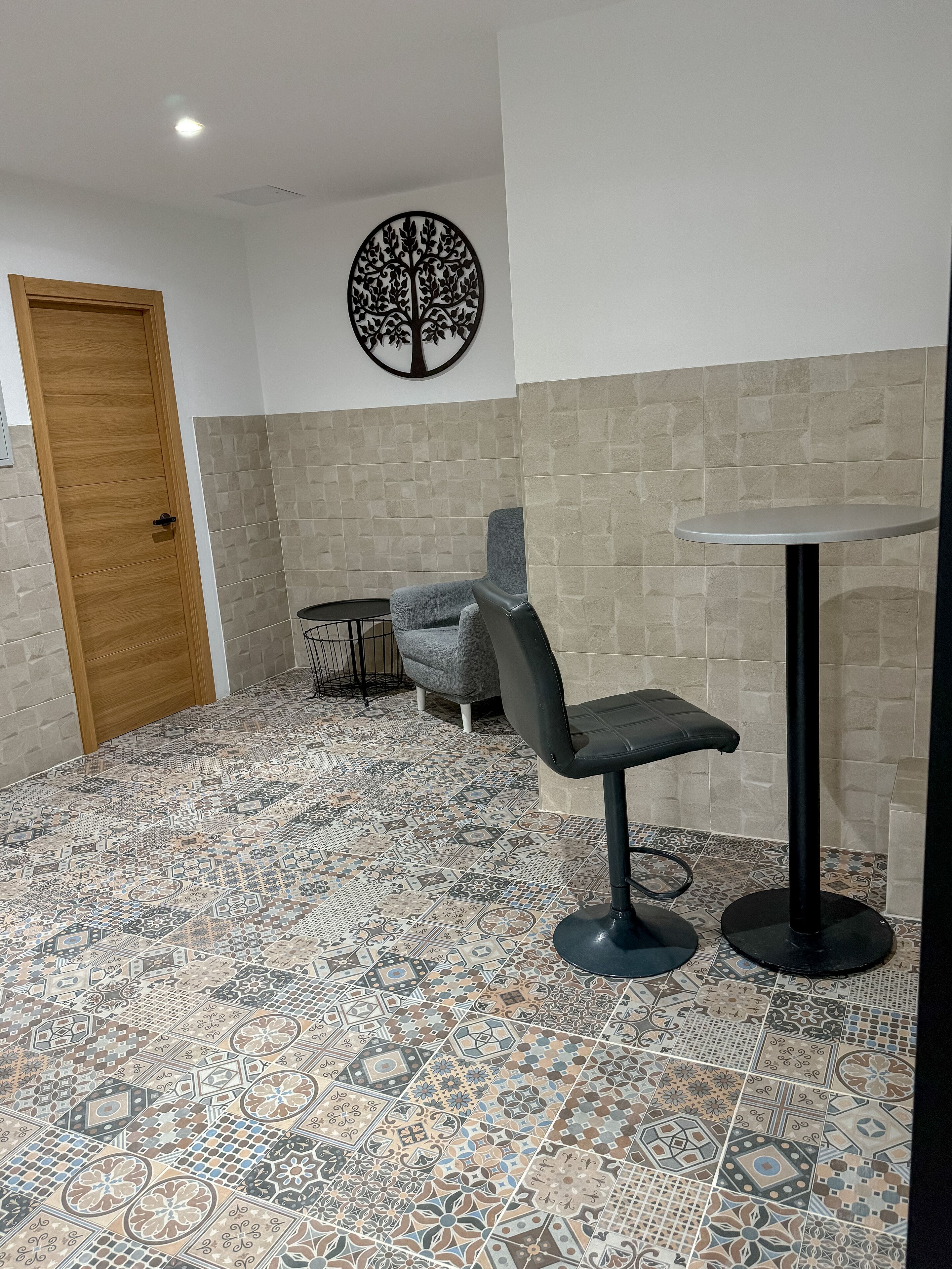 Une pièce avec des carreaux de sol à motifs, une porte en bois, une chaise grise, une table ronde et une décoration en forme d’arbre fixée au mur.