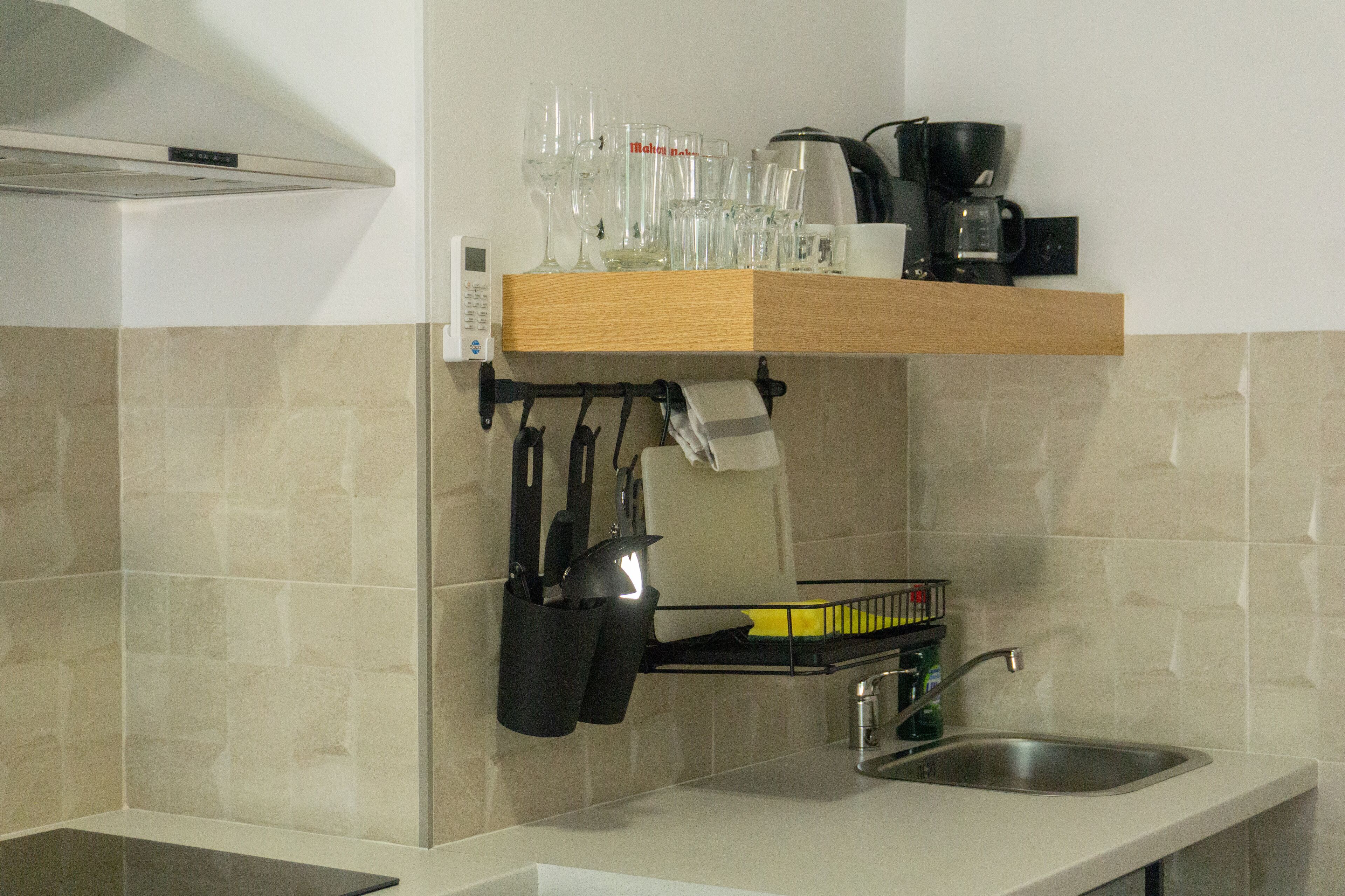 A kitchen with a wooden shelf holding glasses and a coffee maker, a hanging knife rack, and a dish rack.