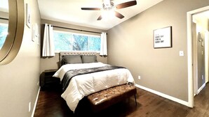 Un dormitorio con cama, ventilador de techo, ventana con cortinas, una cita enmarcada en la pared y una puerta.
