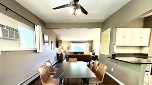 Un comedor con una mesa larga y sillas, un ventilador de techo y una zona de cocina con armarios y encimera.