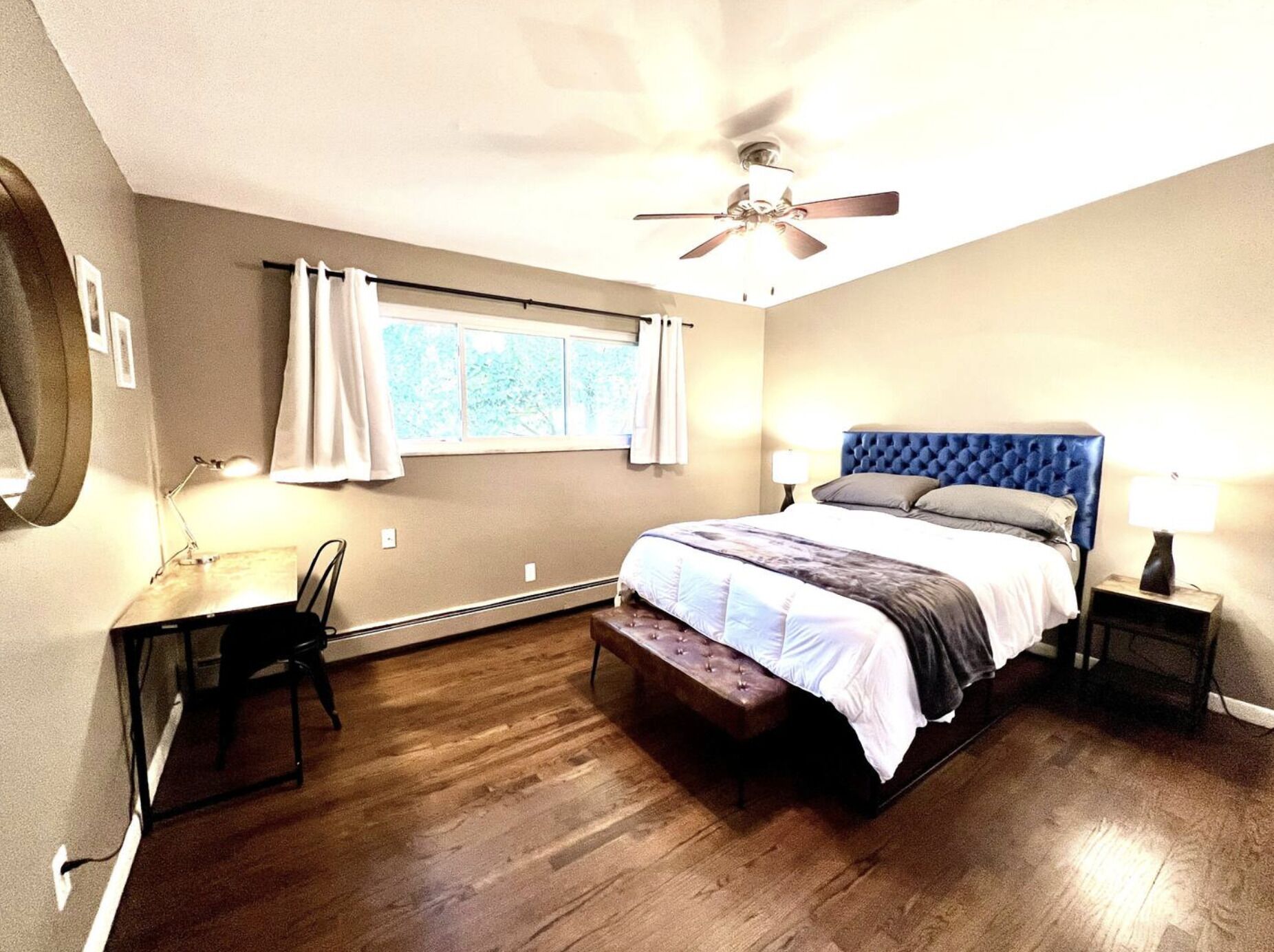A bedroom with a bed, a desk, a chair, a ceiling fan, and a window with curtains.