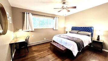 A bedroom with a bed, a desk, a chair, a ceiling fan, and a window with curtains.