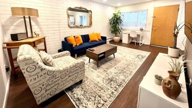 A modern living room with a sofa, coffee table, and a patterned armchair. There's a desk with a lamp, a mirror, and a potted plant. - Stylish 2BR in Historic WaterHill / Walk to Downtown & The Big House (Ann Arbor)