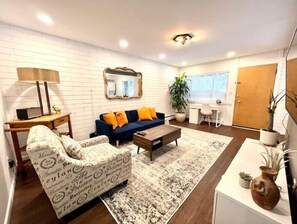 Uma sala de estar moderna com um sofá, mesa de centro e uma poltrona estampada. Há uma escrivaninha com abajur, espelho e um vaso com planta.