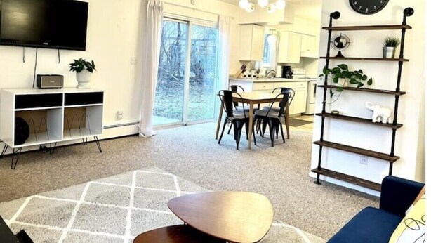 A modern living room with a TV, a dining area with chairs, a kitchen in the background, and a large window.