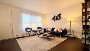 A living room with a dark wooden floor, a large rug, a sofa with colorful cushions, two black chairs, a coffee table, a floor lamp, and a bookshelf.