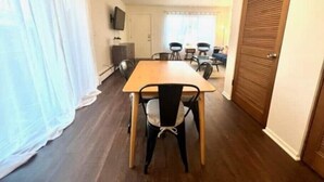 A room with a wooden dining table and chairs, a television on the wall, and a door with a wooden shutter.