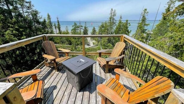 A wooden deck with four Adirondack chairs and a table, overlooking a beach and ocean.