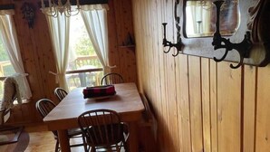 A wooden cabin interior with a dining table, chairs, a chandelier, and a window with curtains.