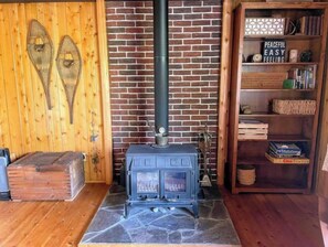 Une pièce rustique avec un mur en briques, un plancher en bois, un foyer en pierre, un poêle ancien et une bibliothèque contenant divers objets.