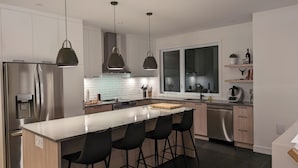 A modern kitchen with stainless steel appliances, a large island with bar stools, pendant lighting, and a view through a large window.