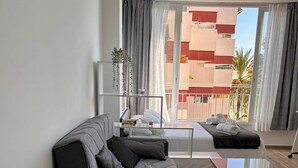 A modern living room with a sofa, a glass coffee table, and a view of a building with balconies. - Lux Studio Nerja (Nerja)
