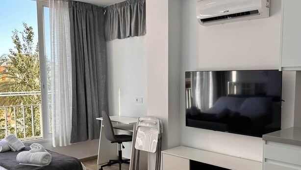 A modern hotel room with a bed, a desk, a flat-screen TV, and a balcony with curtains. - Lux Studio Nerja (Nerja)