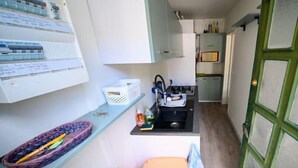 Cocina compacta con un lavamanos, una pequeña encimera y un microondas. Se observa una puerta de marco verde y una cesta blanca sobre la encimera.