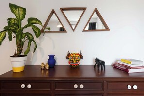 A wooden dresser with three mirrors, a potted plant, a vase, a decorative mask, a small figurine, and a stack of books.