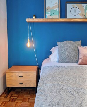 A bedroom with a bed, a nightstand, a wall-mounted shelf, and a hanging light fixture.