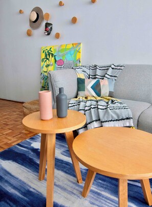 A living room with a grey sofa, two round wooden tables, a blue patterned rug, and a colorful abstract painting on the wall.