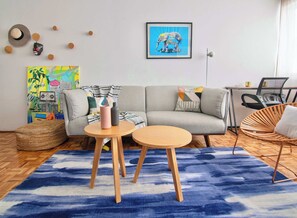 A modern living room with a grey sofa, two round wooden tables, a blue patterned rug, and a framed elephant artwork on the wall.