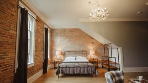 A loft bedroom with exposed brick walls, a sloped ceiling, a bed with a metal frame, two bedside tables with lamps, a chandelier, and a small seating area with a chair and a table.