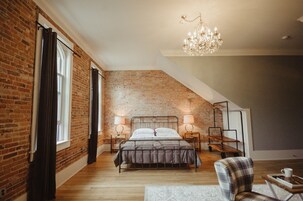 Un dormitorio tipo loft con paredes de ladrillo visto, techo inclinado, cama con estructura metálica, dos mesitas de noche con lámparas, un candelabro y una pequeña zona de estar con una silla y una mesa.