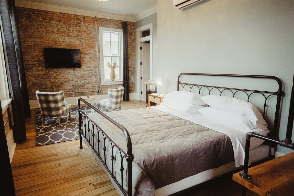 Un dormitorio con estructura de cama metálica, suelo de madera, pared de ladrillo, ventana con cortinas y televisor de pantalla plana montado en la pared.