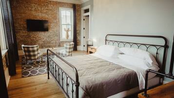 A bedroom with a metal bed frame, a wooden floor, a brick wall, a window with curtains, and a flat-screen TV mounted on the wall.