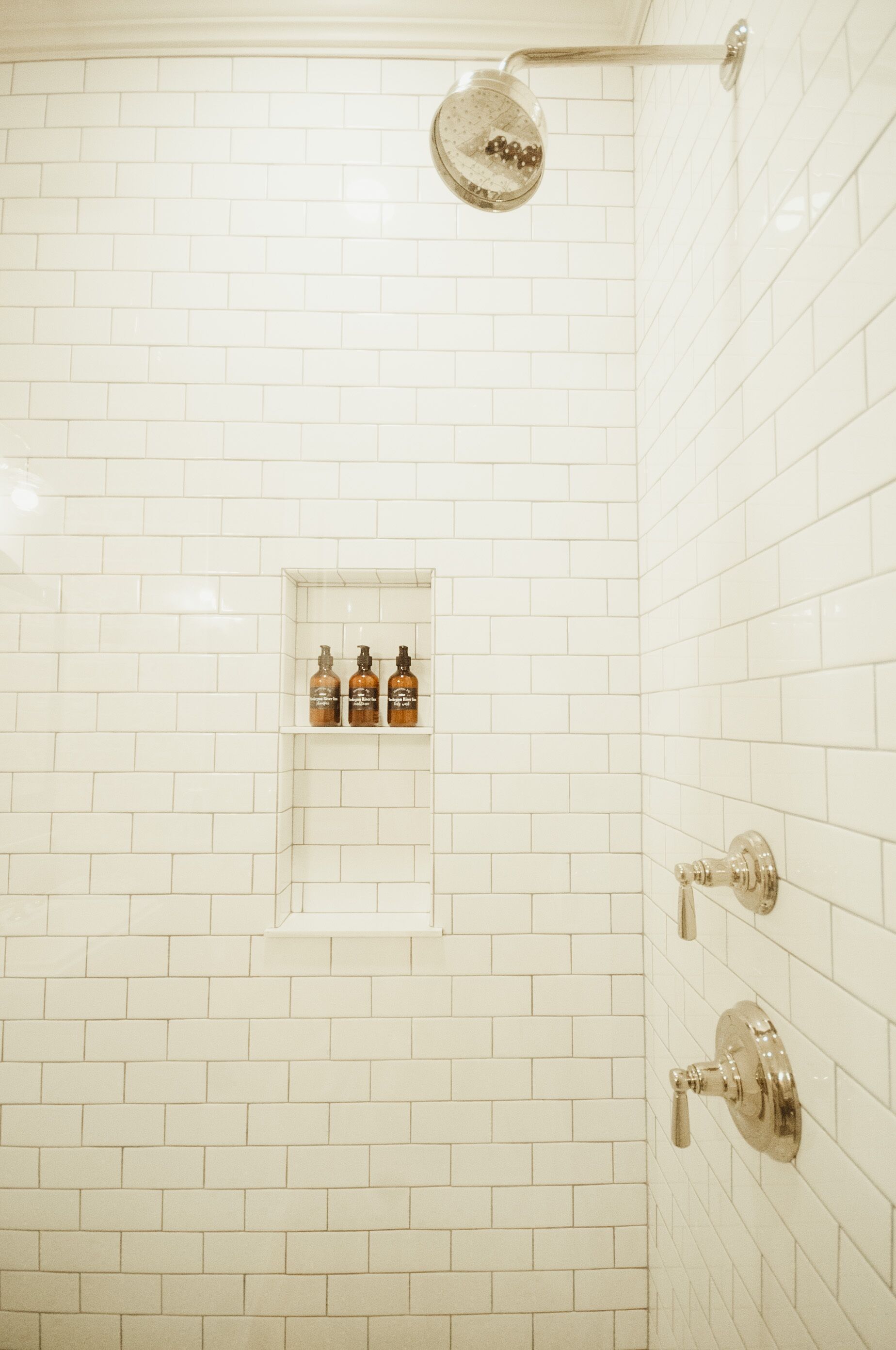 Ducha con azulejos blancos, un estante con botellas y un cabezal de ducha.