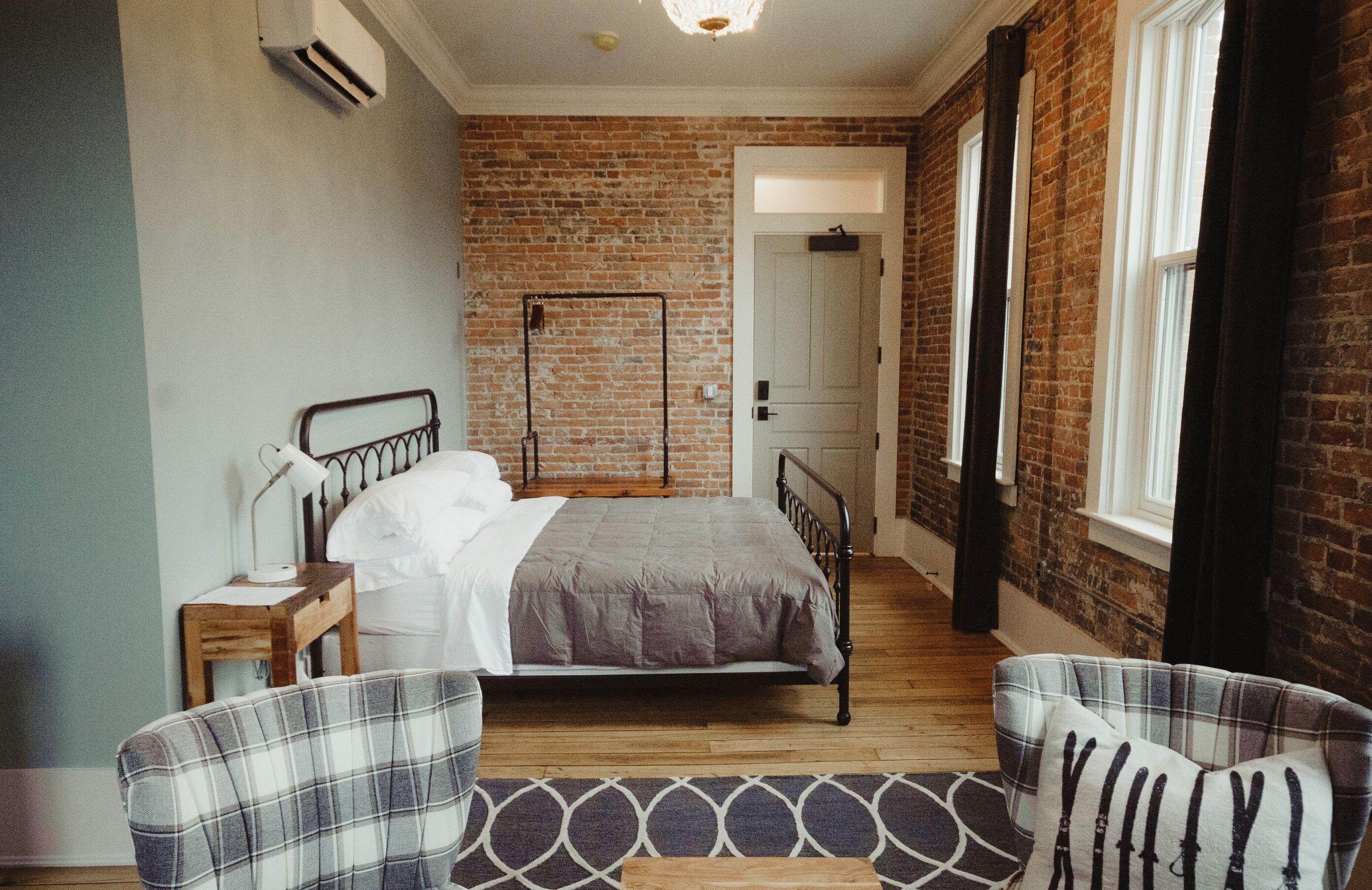 Un dormitorio con pared de ladrillo, una cama, una mesita de noche, una lámpara y dos sillones a cuadros.