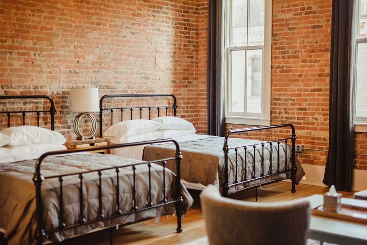 Una habitación con tres camas de metal, una mesita de noche de madera, una lámpara y una ventana con cortinas.