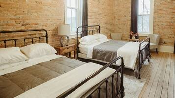 A hotel room with three beds, a chandelier, brick walls, wooden floors, and a window with curtains.