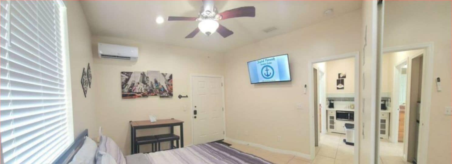 Une chambre à coucher comprenant un lit, un ventilateur de plafond, une télévision et une porte donnant sur la cuisine.