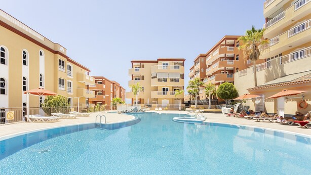 Una piscina rodeada de edificios de varios pisos con balcones, tumbonas y sombrillas.