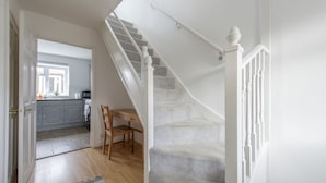 A staircase with white balusters leading to a landing, a wooden chair, and a kitchen area with grey cabinets and a window. - Relax in comfort in Chingford! (Greater London)