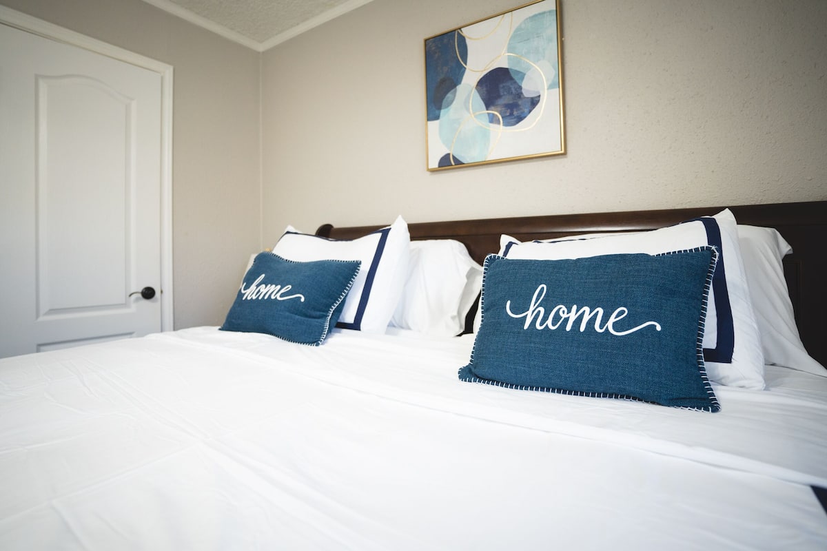 A hotel room with a neatly made bed, two pillows with the word "home" on them, a door, and a wall with an abstract painting.