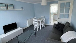 A living room with a grey sofa, a glass coffee table, a white TV stand, a dining table with white chairs, and a door leading to another room. - Peaceful centrally located, 2- Bedroom Bungalow. (Pengegon)