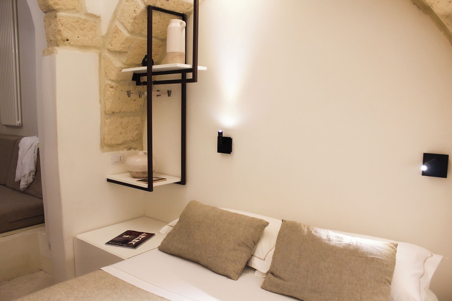 A bedroom with a bed, a nightstand, a shelf with a vase, and a wall-mounted light.