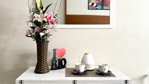Un pequeño comedor con una mesa blanca, dos sillas, un florero de mimbre con flores, una cafetera y una obra de arte enmarcada en la pared.
