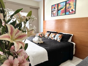 A hotel room with a bed, pillows, a vase of flowers, and a painting on the wall.