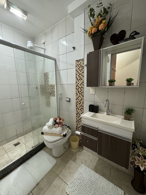 A modern bathroom with a glass shower enclosure, a toilet with a towel and flowers, a sink with a mirror above, and a tiled backsplash.