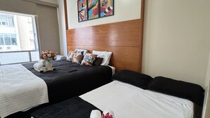 Habitación de hotel con dos camas, ventilador de techo, ventana con vistas y pared decorada con arte colorido.