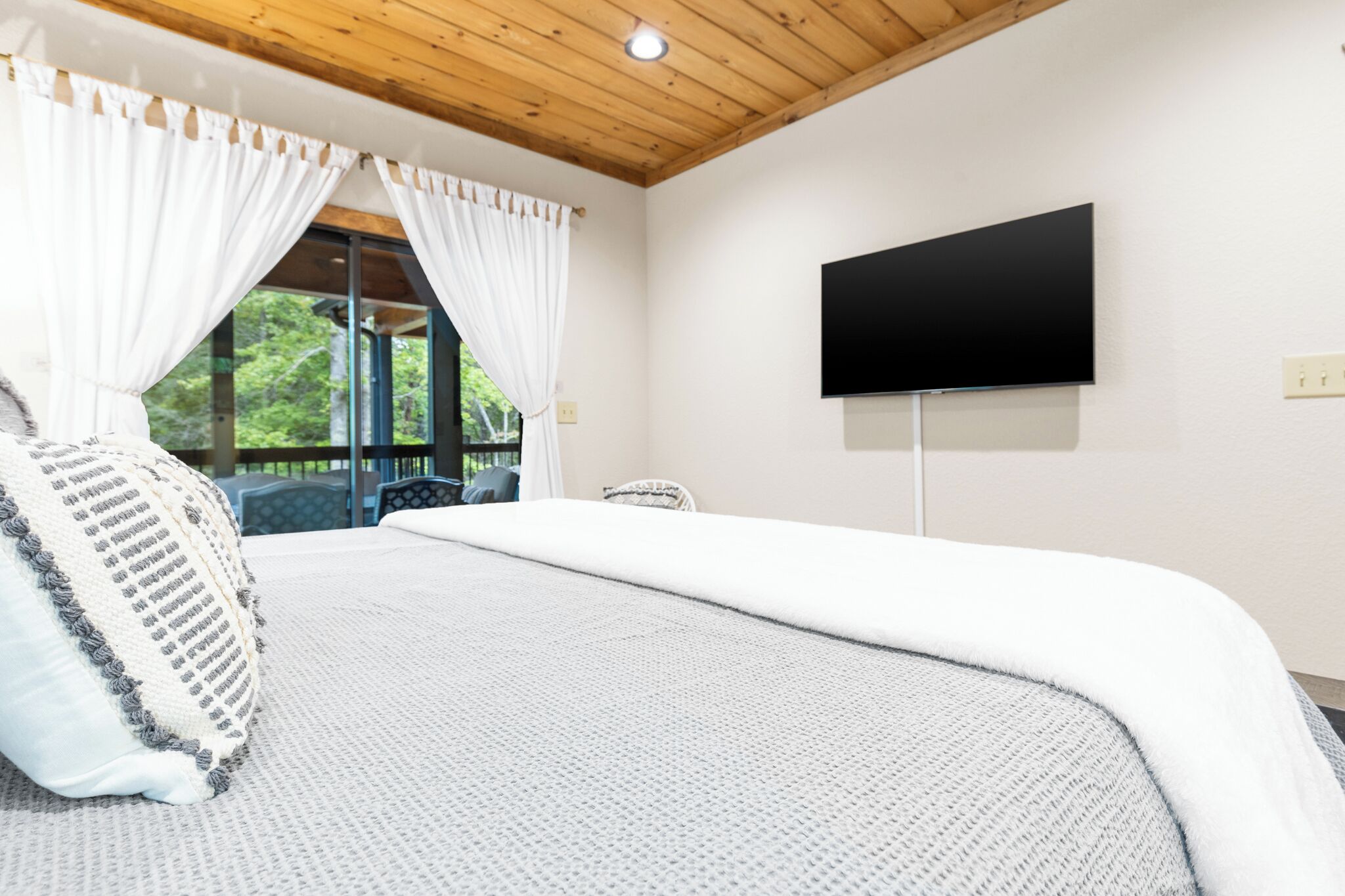 Un dormitorio con una cama grande, cortinas blancas, un televisor de pantalla plana en la pared y vistas a zonas verdes al exterior.