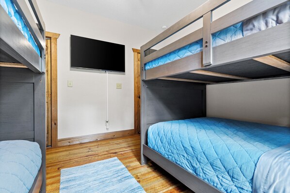 Habitación con literas, un televisor montado en la pared, piso de madera y colcha azul acolchada.