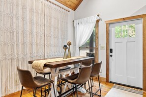 A dining area with a wooden table, four chairs, a macrame wall, a white door, and a window with a view of trees. - Hidden Haven- Welcome to Modern Living! (Morganton)