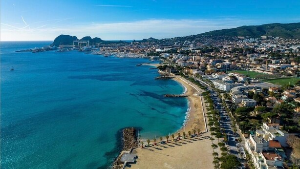 Une vue aérienne d’une ville côtière, avec une plage, des eaux bleues et limpides, et une zone urbaine dense, avec des bâtiments et des routes.