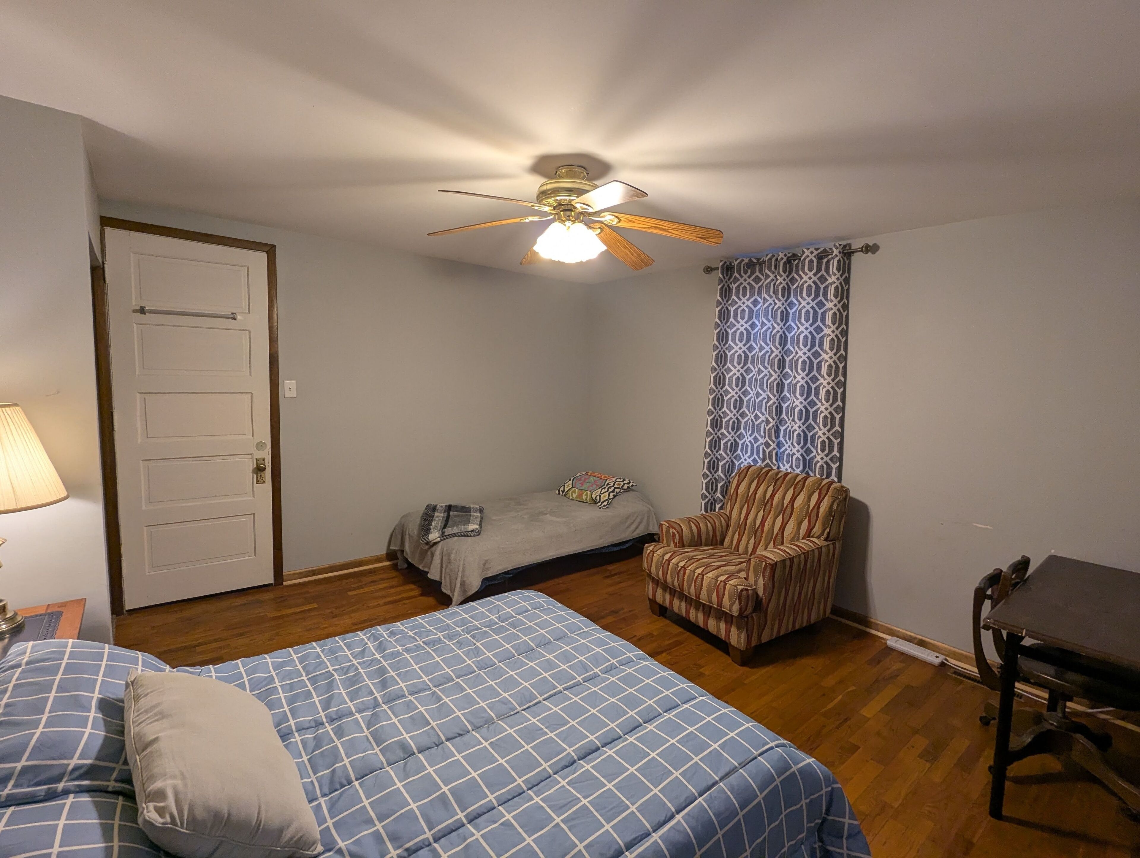 A room with two beds, a chair, a ceiling fan, a patterned curtain, and a door.