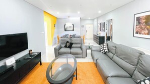 Een moderne woonkamer met een flatscreen-tv, grijze banken, een glazen salontafel en een eethoek op de achtergrond.