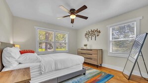 A bedroom with a bed, a ceiling fan, a dresser, a window with blinds, and a standing desk.